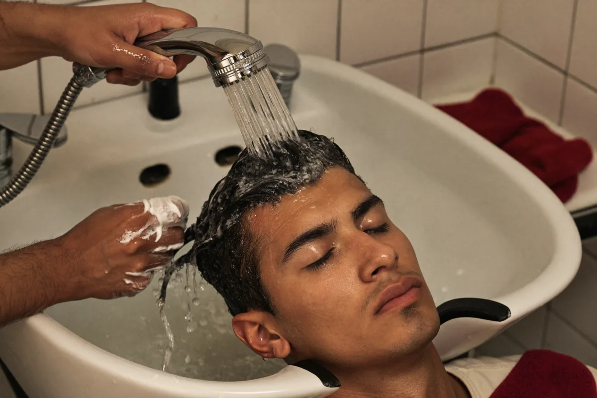 A barber rinsing shampoo lather from a young man’s hair over a salon basin with a handheld shower
